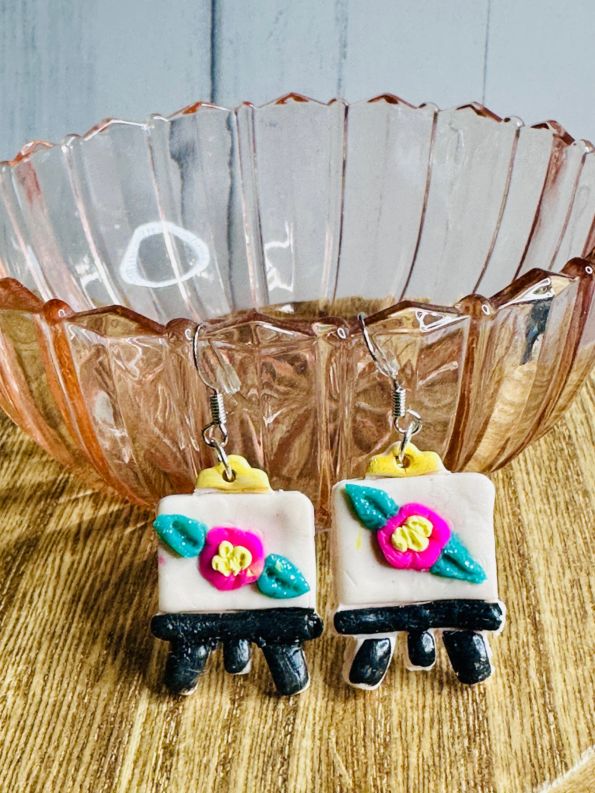 A pair of Art Canvas with Flower Earrings leaning against a pink glass bowl.  Each earring has a silver hoop that connects to the top of the art canvas.  The canvas is white with a pink, green, and yellow flower.  The feet of the canvas are black.