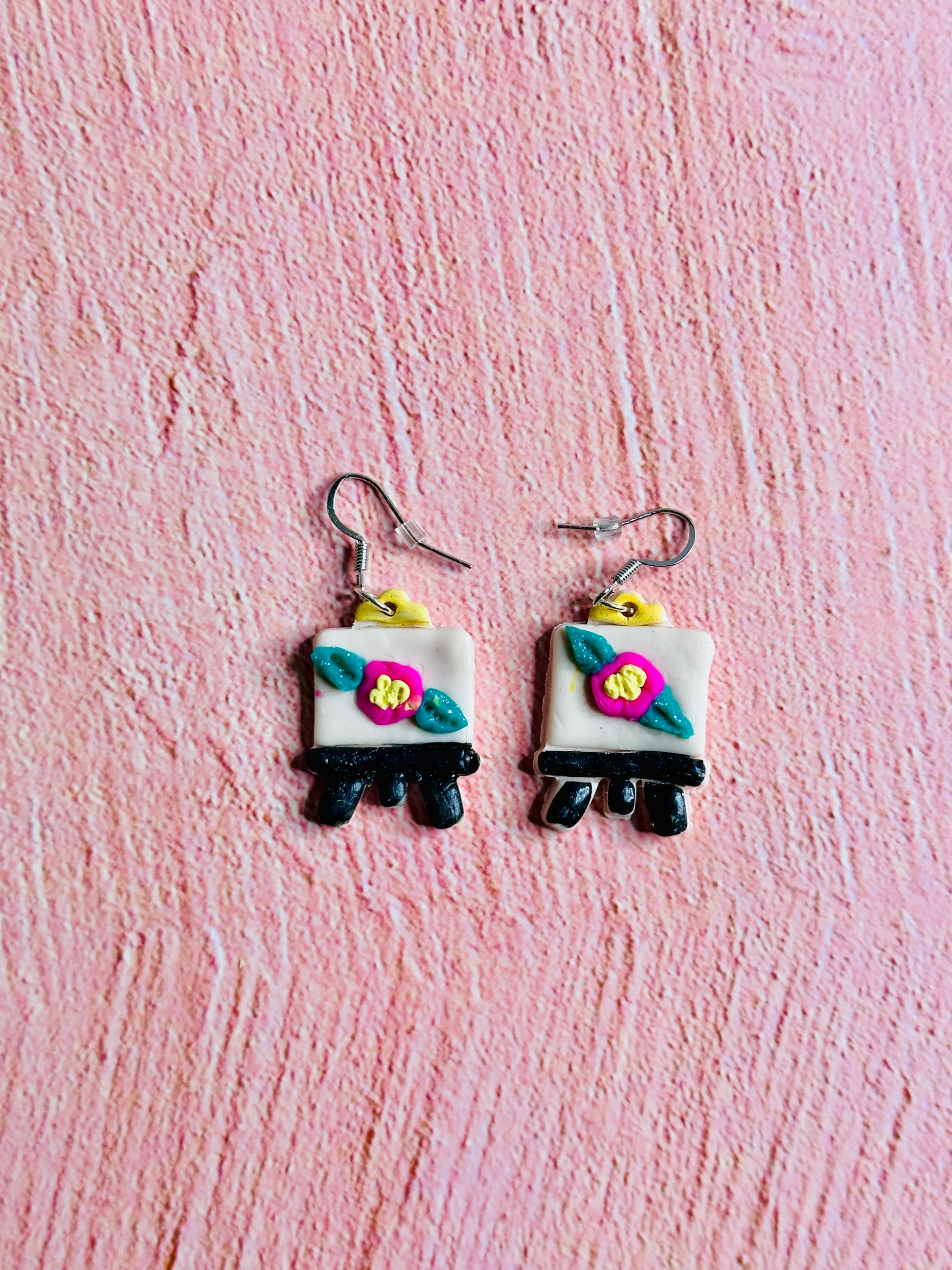 A full-shot image of two Art Canvas with Flower Earrings on a pink background. Each earring has a silver hoop that connects to the top of the art canvas.  The canvas is white with a pink, green, and yellow flower.  The feet of the canvas are black.