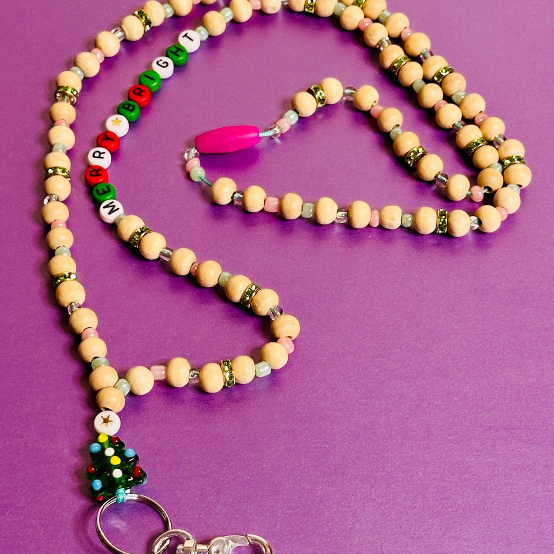 The Christmas Tree Lanyard viewed from an angle with the Christmas Tree charm on the bottom left, the beaded words "MERRY AND BRIGHT" on the upper left, and the pink breakaway clasp in the middle.  The lanyard is laying in a circular shape.