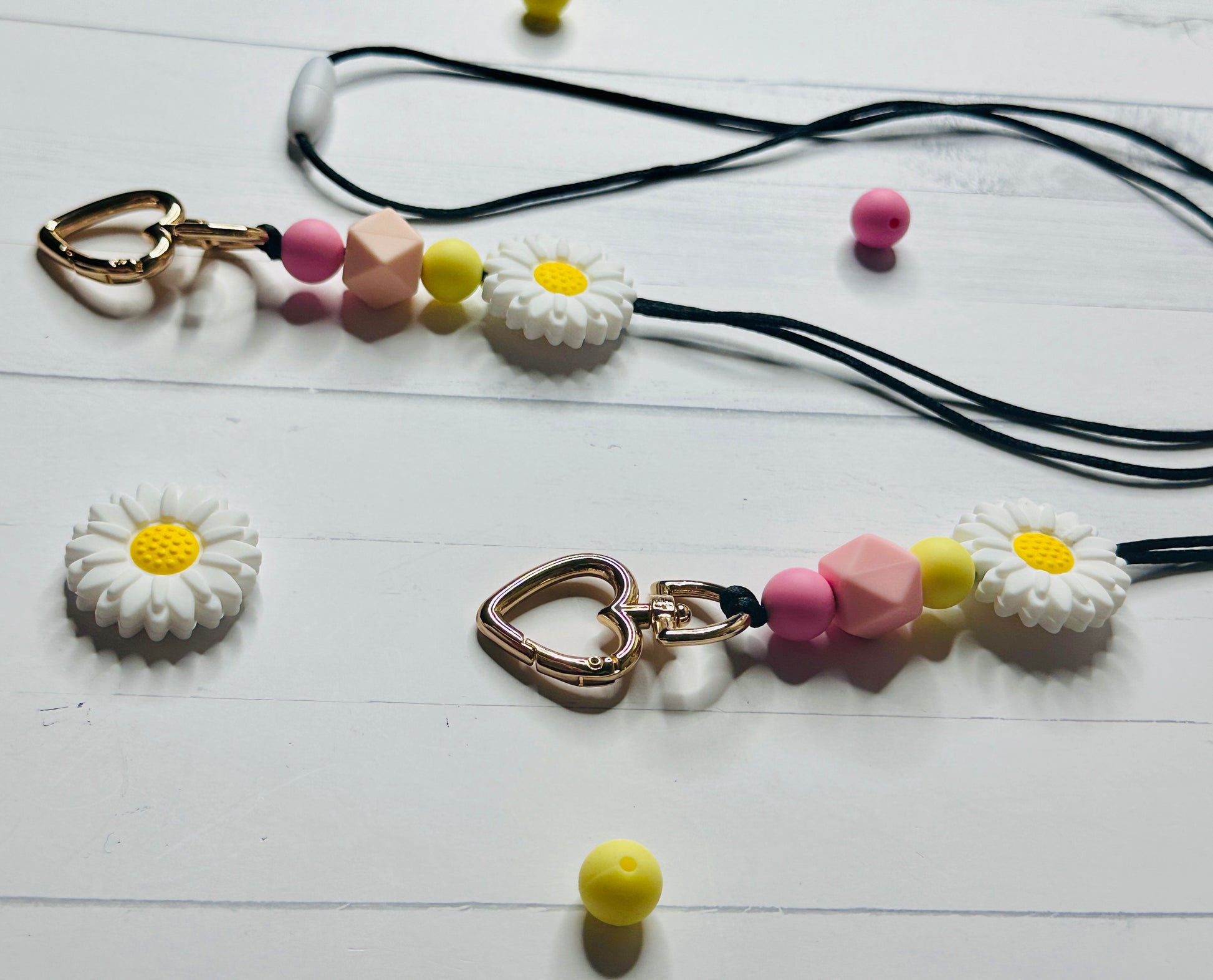 A close up and angled view of the Flower Lanyard.  The heart shaped metal clasp, silicon beads, and daisy flower are displayed on a white wooden table and are casting shadows onto its surface.