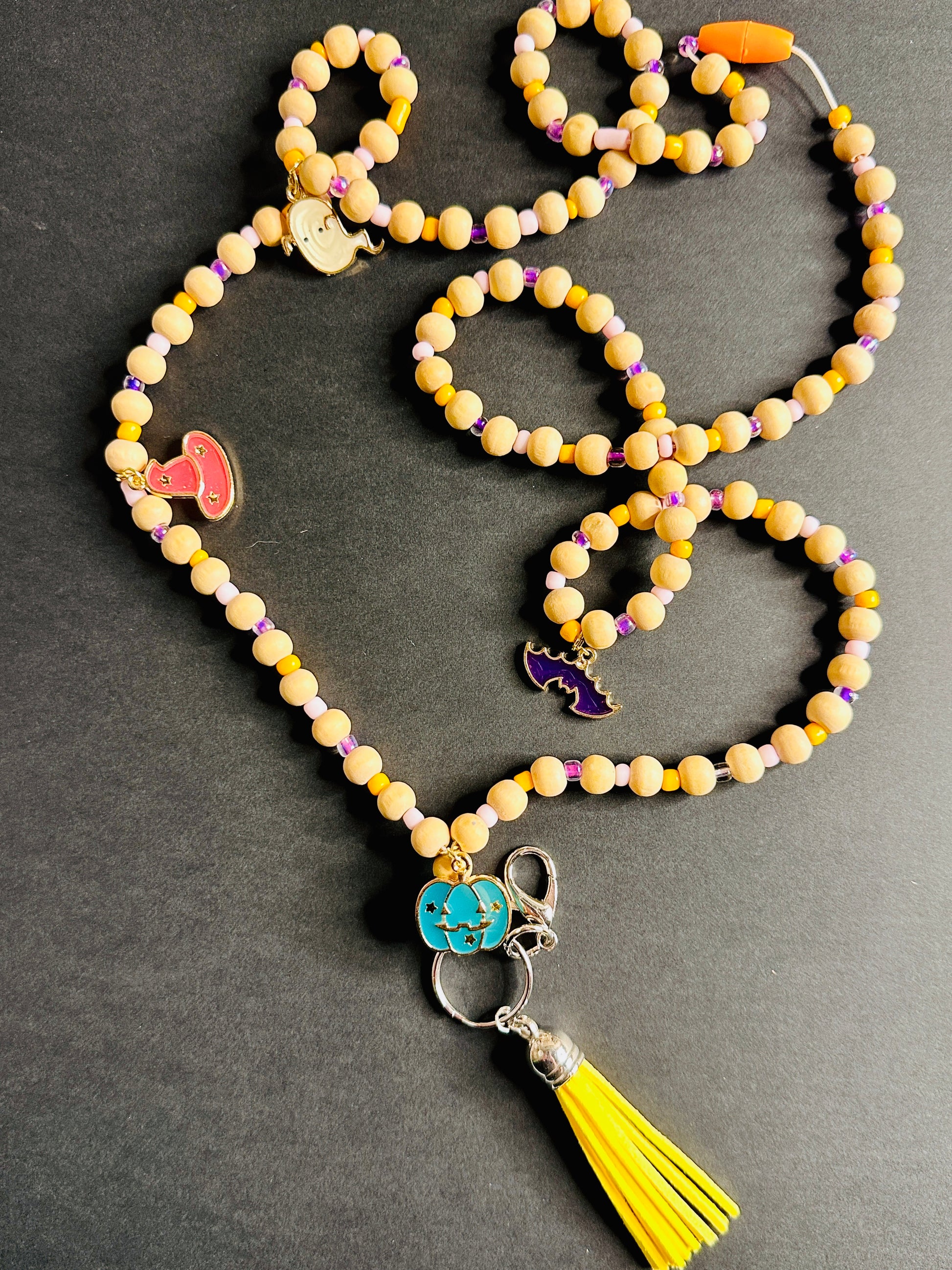 The Halloween Lanyard on a grey background.  The full lanyard can be seen with its wooden beads, metal charms shaped like pumpkins, bats, witch hats, and ghosts, and colorful tassel.